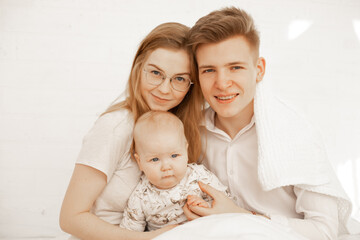 Fototapeta na wymiar Happy young family of three people sit together and embrace, white background, free copy space. Home family photo of mom, dad and infant child. Concept of parental affection and caring for children