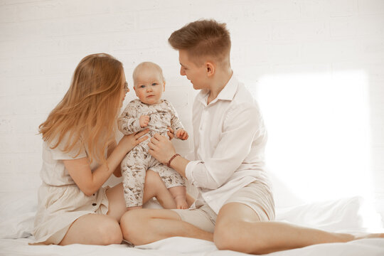 Happy young couple hold in hands little baby and have fun sitting on bed, white background, free copy space. Home family photo of mom, dad and infant child. Parental affection and love