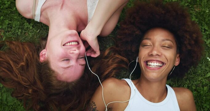 Portrait of Two Multiethnic Women Lying Down on a Park Lawn Using Earbuds to Listen to Music. Black Female Teenager Sharing her Favorite Song with her Friend While Tey are Sunbathing on a Hot Day