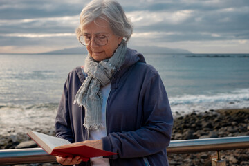 Relaxed senior woman in retirement at sea reading a book at sunset - atlantic ocean in Tenerife, canary islands. Travel and freedom concept