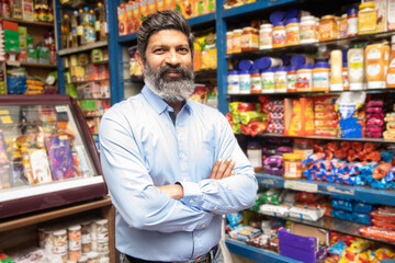 Portrait of happy mature Indian man standing at grocery shop or supermarket with cross arm. - obrazy, fototapety, plakaty