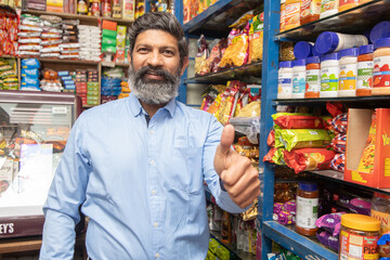 Portrait of happy mature Indian man standing at grocery shop or supermarket do thumbs with his hand. Small business owner