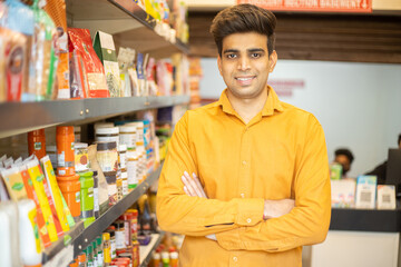 Portrait of happy handsome young Indian man wearing orange shirt standing cross arm at grocery shop or supermarket, Closeup. Selective Focus.
