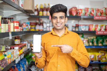 Portrait of happy handsome young Indian man showing smart phone with blank display screen to put advertisement at grocery shop or supermarket, Closeup.