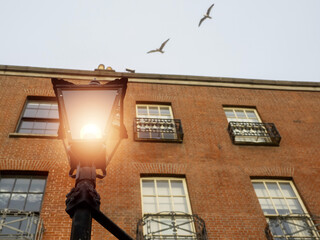 Fototapeta na wymiar Old style lantern lamp in a street. Old red brick building in the background. Sea gull flying high in the air out of focus. City illumination. Warm light glow.