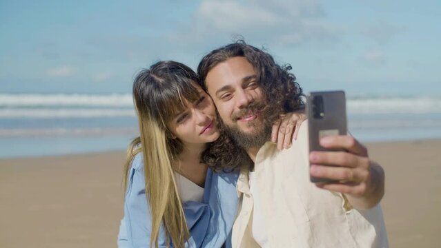 Beautiful couple taking selfie at seashore on sunny day. Bearded guy with long wavy hair and pretty blonde girl hugging while man holding phone and kissing girlfriend. Mobile technology, love concept