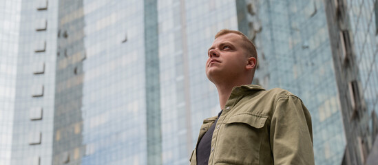 A pensive man stands against the backdrop of a skyscraper.