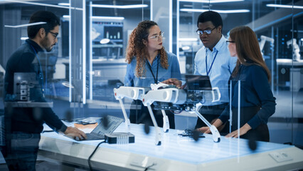 Diverse Team of Industrial Robotics Specialists Gathered Around a Table With a Mobile Robot. Scientists Use Tablet and Laptop Computers to Program the Automated AI Robotic Dog.