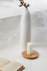 Fototapeta na wymiar Still life photo of a white candle with a wavy surface. The designer handmade candle is located on a wooden board against a light wall. There is a white vase with flowers and a book on the table.