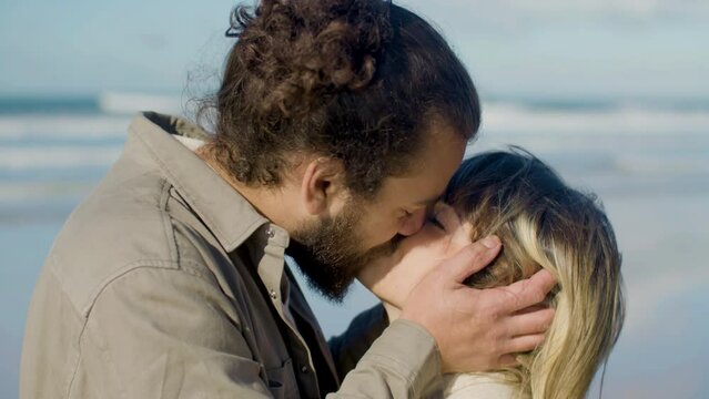 Closeup shot of young Caucasian couple kissing and talking against sea background. Handsome guy with man bun holding girls face in his hands, caressing and hugging. Love, romance concept