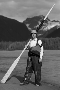A man stands on the shores of a river for a portrait with an oar, wool hat and rain slicker overalls.