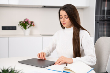  Young woman opens laptop to continue homework. Female student sits at desk in stylish kitchen working on task on modern laptop