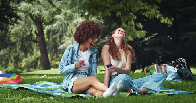 Portrait of Two Multiethnic Female Friends Sitting in a Park, Having a Picnic, Watching Funny Videos on the Internet Using Smartphone. Female Teenagers Sharing Memes and Laughing