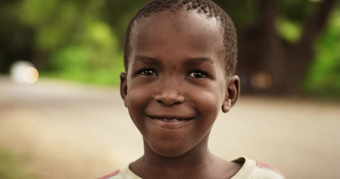 Close Up Portrait of a Shy Authentic African Boy Smiling at the Camera with a Blurred Rural Village in the Background. Black Male Kid Representing Future, Hope, and Acceptance. Documentary Footage