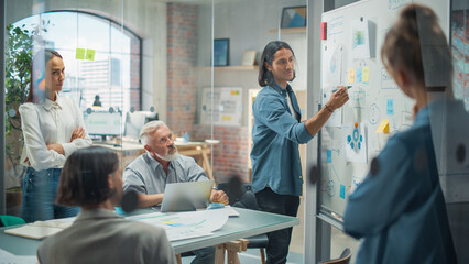 Project Manager Makes a Presentation for a Diverse Creative Team in Meeting Room in an Agency. Colleagues Sit Behind Conference Table and Discuss Business Development, User Interface and Design.