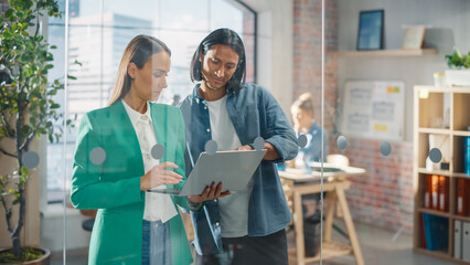 Two Creative Colleagues Discussing and Pointing at a Laptop in Modern Casual Office. Female Caucasian Sales Manager Discussing Project Plan with Male Latin Legal Assistant and Taking Notes