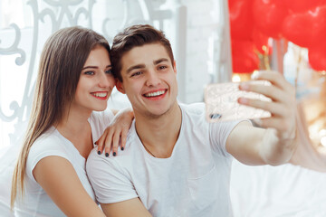 Beautiful young couple taking selfie on mobile phone, sitting at home and wearing white T-shirts, celebrating Valentine's day.