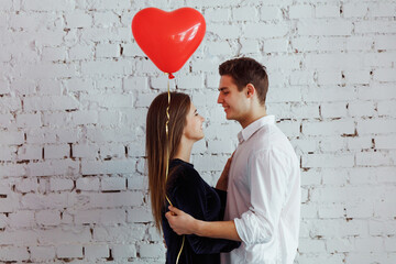 Obraz na płótnie Canvas Beautiful young couple hugging on the white wall background while celebrating Saint Valentine's Day with air balloon in shape of heart in hands.