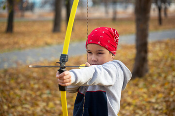 Asian 7 years old boy in a red bandana aim with a bow outdoor in public park in autumn, close up, selective focus.
