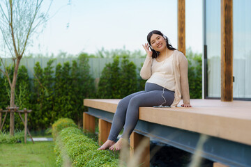 Fototapeta premium Portrait of pregnant Happy Asian woman sitting on terrace at home and using smart phone for listening music in headphones.