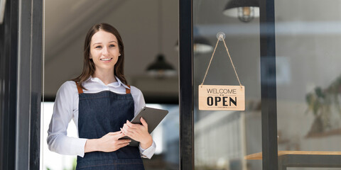 Portrait of a happy waitress standing at restaurant entrance. Portrait of mature business womanattend new customers in her coffee shop. Happy woman owner showing open sign in her small business shop.