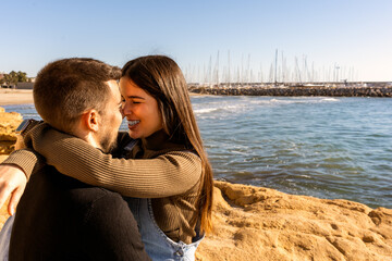 Optimistic young woman in casual clothes hugging and touching noses with man on rocky seashore on Saint Valentine Day