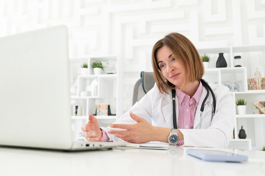 Portrait photo of young and beautiful thinking female doctor in white medical gown sitting and working on laptop in the office of the modern clinic. Online consultation and distant cure concept