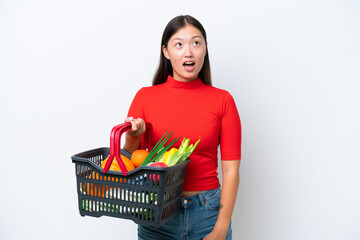 Obraz na płótnie Canvas Young Asian woman holding a shopping basket full of food isolated on white background looking up and with surprised expression