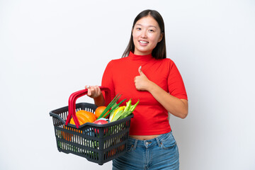 Plakat Young Asian woman holding a shopping basket full of food isolated on white background giving a thumbs up gesture