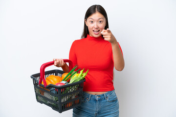 Obraz na płótnie Canvas Young Asian woman holding a shopping basket full of food isolated on white background surprised and pointing front