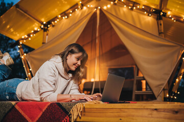 Obraz na płótnie Canvas Happy Woman freelancer using a laptop on a cozy glamping tent in a summer evening. Luxury camping tent for outdoor holiday and vacation. Lifestyle concept