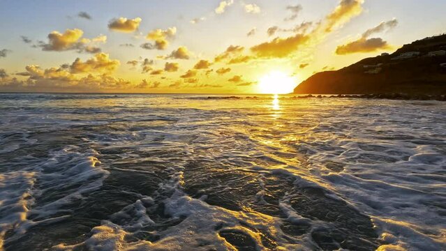 Golden sun dips into the ocean behind headland at Little Bay, St. Maarten - Waves break and roll ashore at camera level