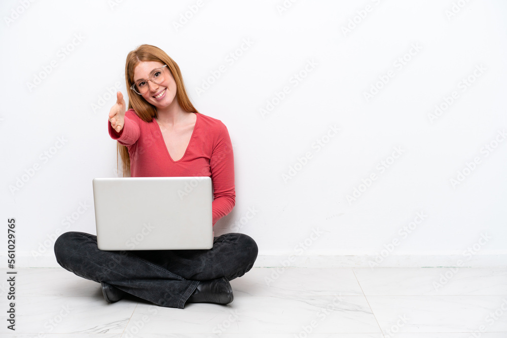 Poster Young redhead woman with a laptop sitting on the floor isolated on white background shaking hands for closing a good deal