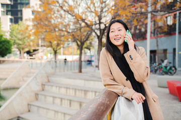 Happy asian young woman having a friendly call conversation using a smartphone standing outdoors. One female smiling and talking by cellphone on the street. Communication concept. High quality photo