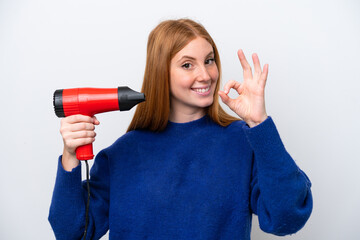 Young redhead woman holding a hairdryer isolated on white background showing ok sign with fingers