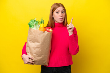 Young redhead woman holding a grocery shopping bag isolated on yellow background intending to realizes the solution while lifting a finger up