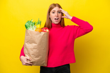 Young redhead woman holding a grocery shopping bag isolated on yellow background with surprise...