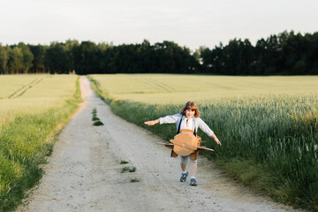 A cheerful kid in aviator's glasses runs in a cardboard plane. Wheat field background lit by sunlight. Children's games and dreams. High quality photo