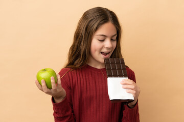 Little caucasian girl isolated on beige background taking a chocolate tablet in one hand and an...