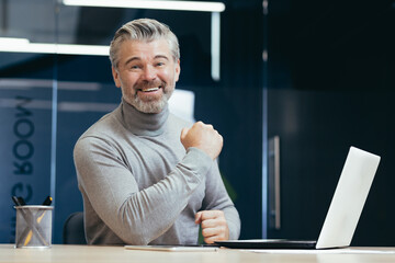 Portrait of a senior gray-haired successful businessman. A happy man is sitting in the office at the table with a laptop. Celebrates victory, success, smiles at the camera, shows well done.