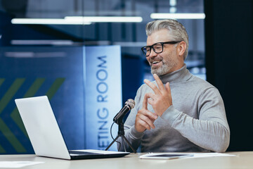 Senior gray-haired man sitting at a desk in an office with a microphone and laptop. Talks on a video call. Conducts interviews, business meetings, webinars, online meetings. Shows, counts on fingers.