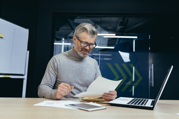 Senior gray-haired man working in office at desk with laptop and documents. an envelope with a letter, bills, and reports, good news.
