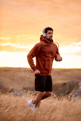 Muscular athletic male in hoodie training jogging outdoors, listening to music, on field, fit caucasian brunette guy in headphones, running alone. sport, fitness, healthy lifestyle concept.