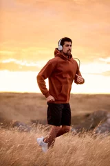 Poster Muscular athletic male in hoodie training jogging outdoors, listening to music, on field, fit caucasian brunette guy in headphones, running alone. sport, fitness, healthy lifestyle concept. © Roman