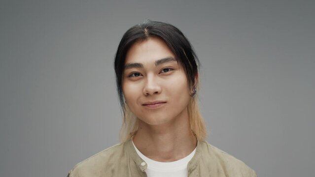Young Adult Korean Man Looking at Camera Smiling Close-up. Portrait of Japanese Boy Isolated Alone on Grey Background. Asian 20s Person with Beautiful Eyes Turning Head and Happy in Colour Studio Shot