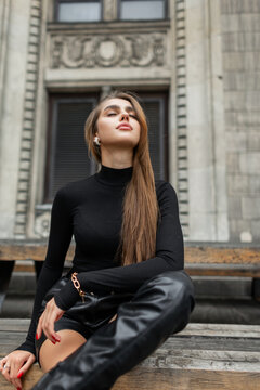 Beautiful glamorous trendy young woman in fashion black clothes with long sleeve golf sweater, leather shorts and high boots is sitting and rest near the wine building