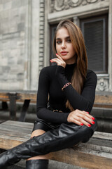 Cool fashionable woman model in fashionable black summer outfit with long sleeve golf, shorts and high shoes sits on a wooden bench near a vintage building