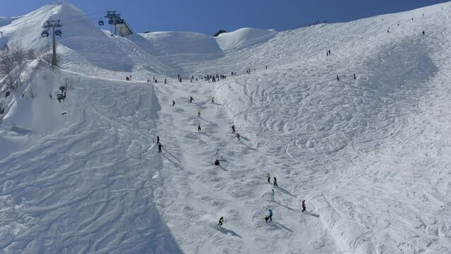 Aerial raising view tourists people riding ski snowboard on slope track at mountain valley resort ropeway cabins. Drone shot extreme sport recreation adrenaline hobby leisure activity winter sunny day