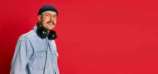 Portrait of man in stylish modern clothes, glasses, hat and headphones posing over red background. Banner, flyer. Concept of modern fashion, lifestyle, music culture
