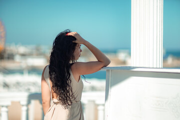 Sea woman rest. A woman with long curly hair in a beige dress stands with her back and looks at the sea and the coast from a balcony with balusters. Tourist trip to the sea.